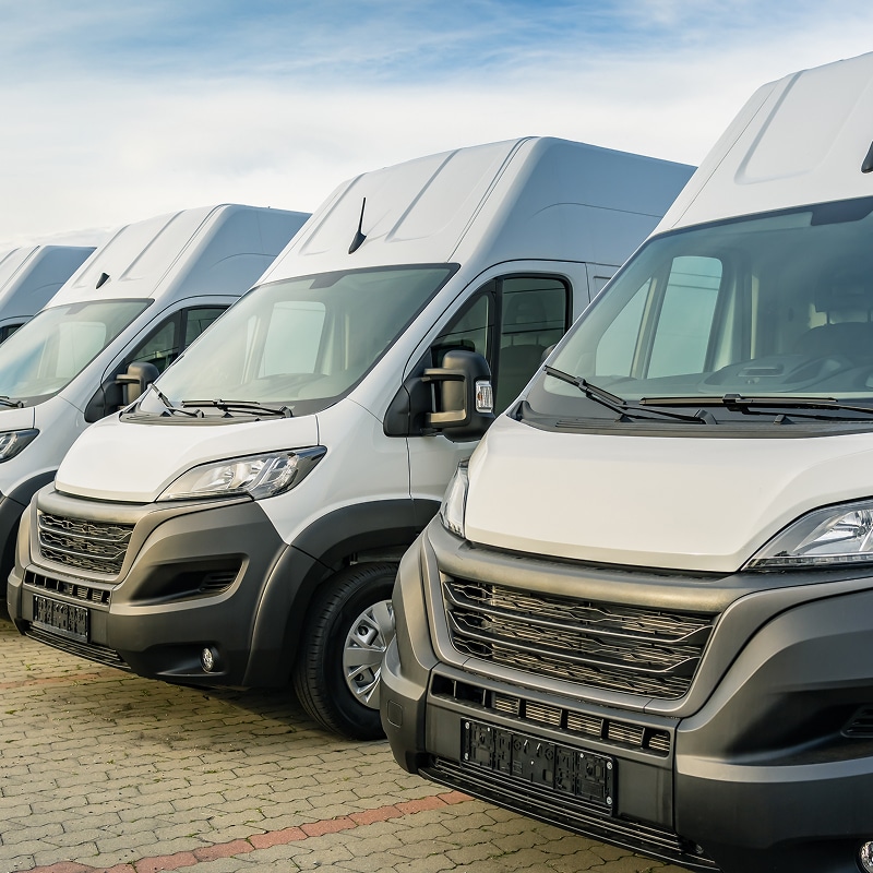 Fourgons Utilitaires Blancs Neufs – Achat ou Location Flotte Flotte de fourgons utilitaires blancs neufs à toit haut, alignés en rangée sur un parking en pavés. Logistique et livraison.