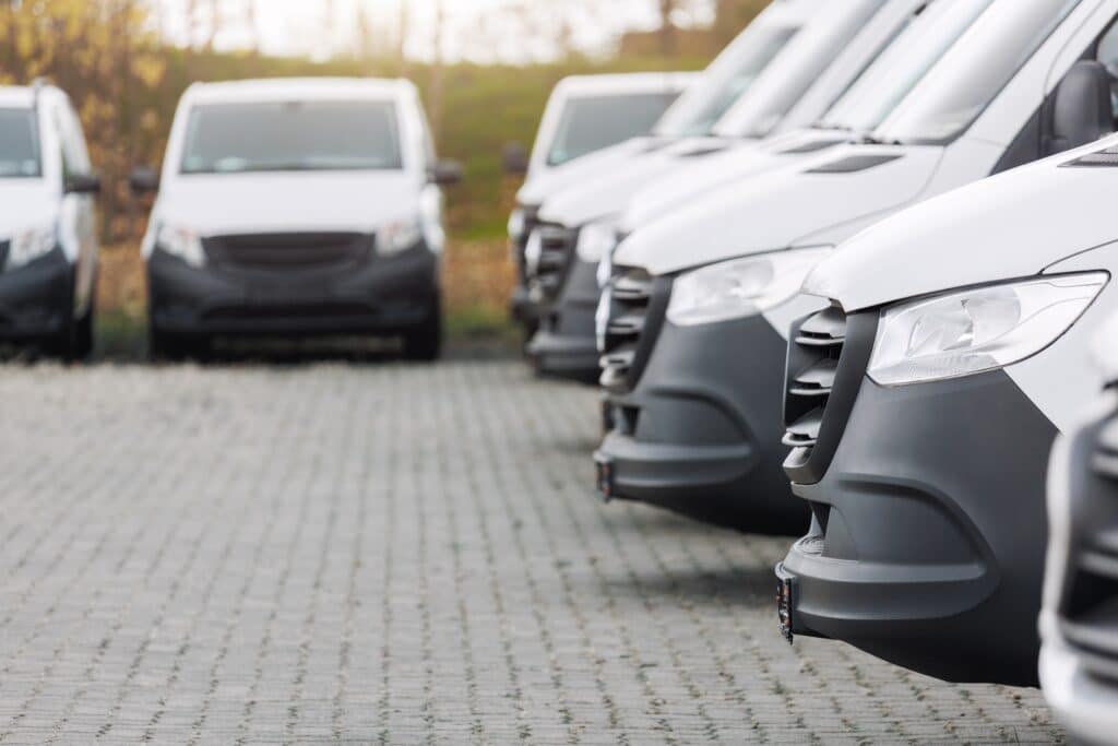 Flotte de vans utilitaires blancs neufs alignés. Vue en gros plan sur les pare-chocs et phares avant sur un parking extérieur.