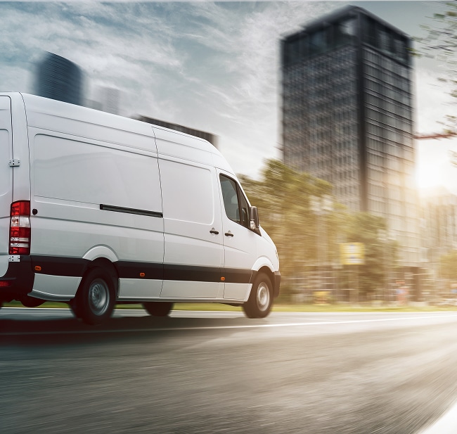 Livraison rapide en ville : fourgon blanc en mouvement Fourgon de livraison blanc roulant rapidement en ville. Mouvement flou sur la route devant des gratte-ciel sombres sous un ciel nuageux et un soleil intense.